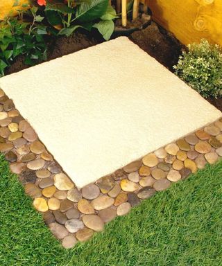 A backyard with a beige square with brown pebbles on the border and a dark green lawn