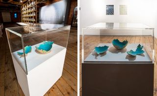 Three broken bowls in pale orange, with the blue and green inside part that bleeds over the uneven edge, sit in an enclosed glass case on a white platform.