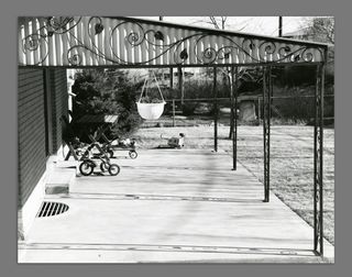 Black and white image of the porch of a house with a Childs trike and toys scattered