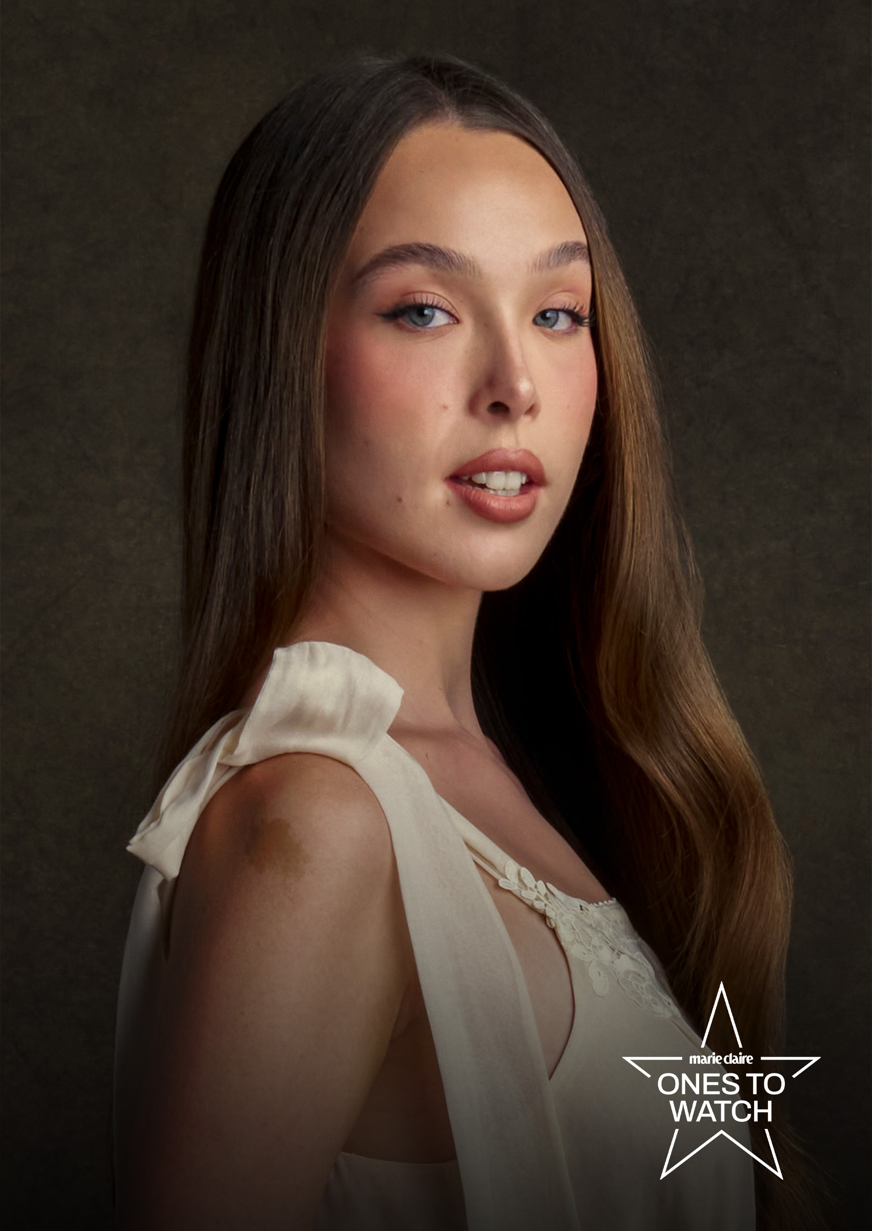 actress ella beatty poses in a portrait looking over her shoulder and wearing a white dress with a ribbon on the shoulder