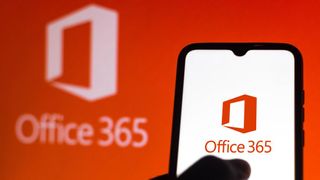 A hand pressing a phone with the Office 365 logo shown on it, with the Office 365 logo on an orange wall in the background