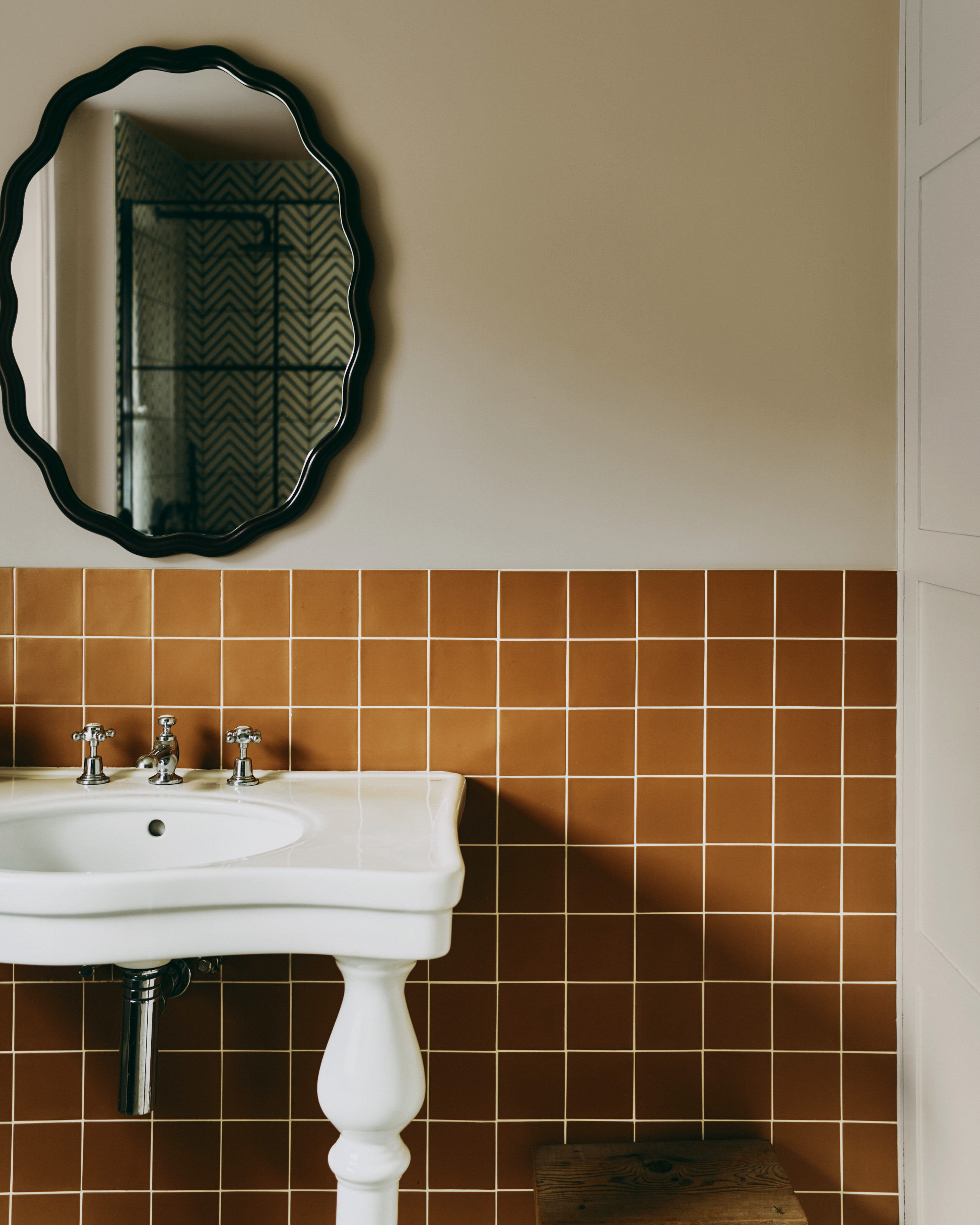 Bathroom with cream walls, terracotta orange square tiles, white traditional basin and black wavy oval mirror