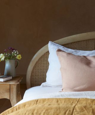 A light brown rattan headboard on a bed with white sheets, a pale pink throw pillow and mustard throw on it, with a wooden nightstand to the left with a silver jug of flowers and a book on it, against a dark brown wall
