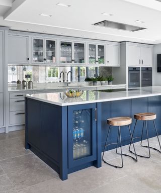 Kitchen with mirror tiled wall, white walls, and dark blue kitchen island with integrated fridge and bar stools.