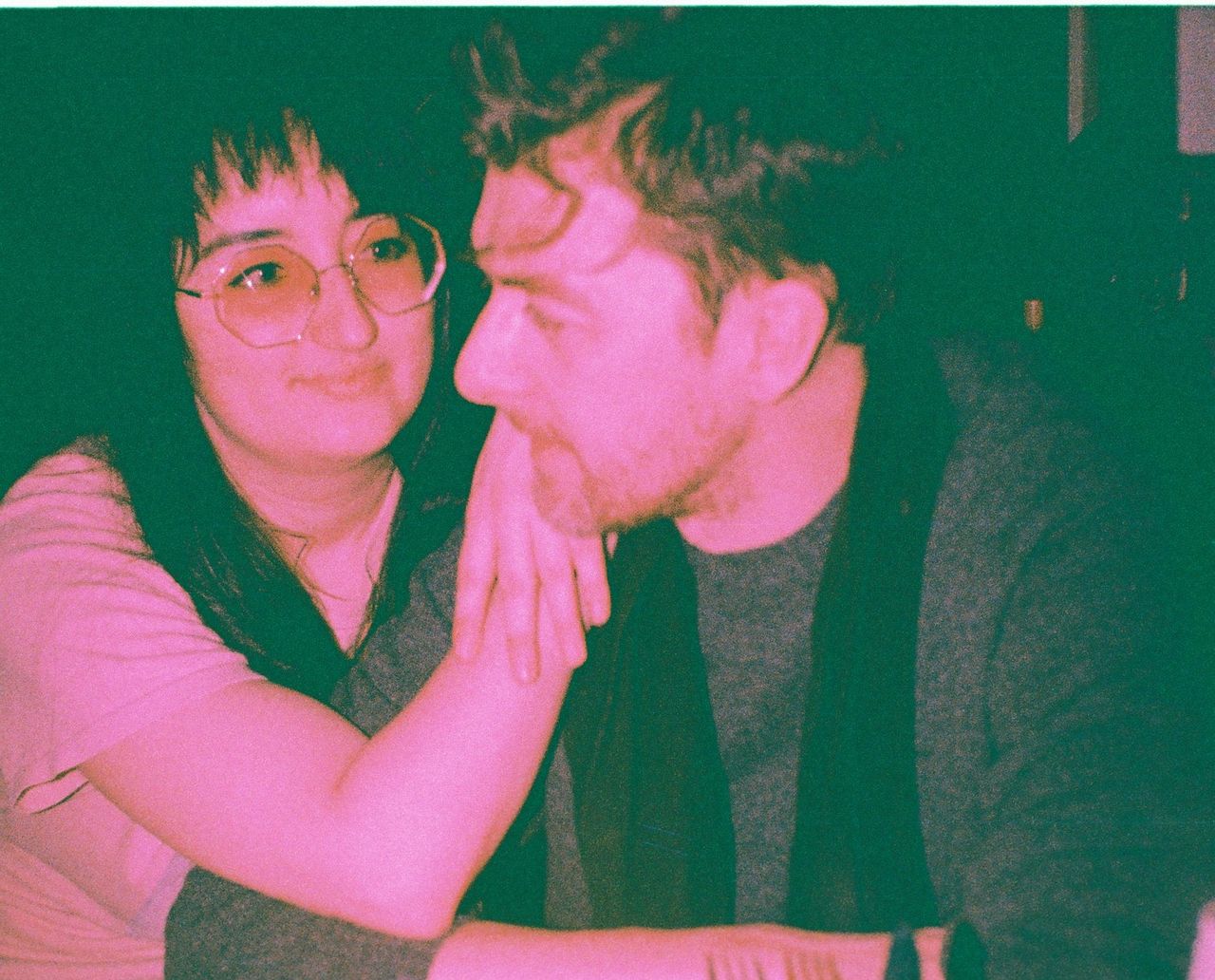 Two people in close conversation; one wearing glasses gently resting their arm on the other's shoulder in a dimly lit setting