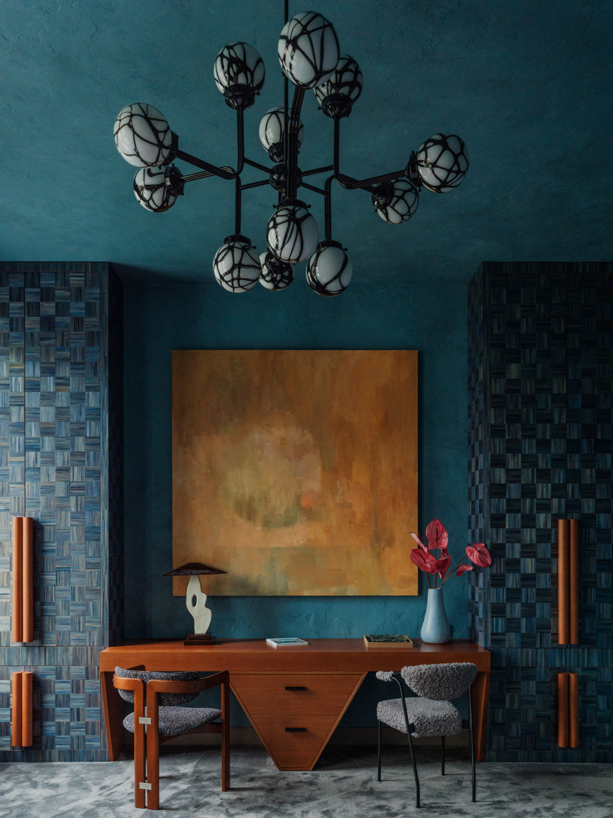 Image of a room that has dark teal walls and ceilings with floor-to-ceiling cabinetry that is a blue-stained wood with a basket weave pattern. There are gray carpeted floors, large orange handles on the cabinets, a large orange painting on the wall, and a geometric wooden desk.