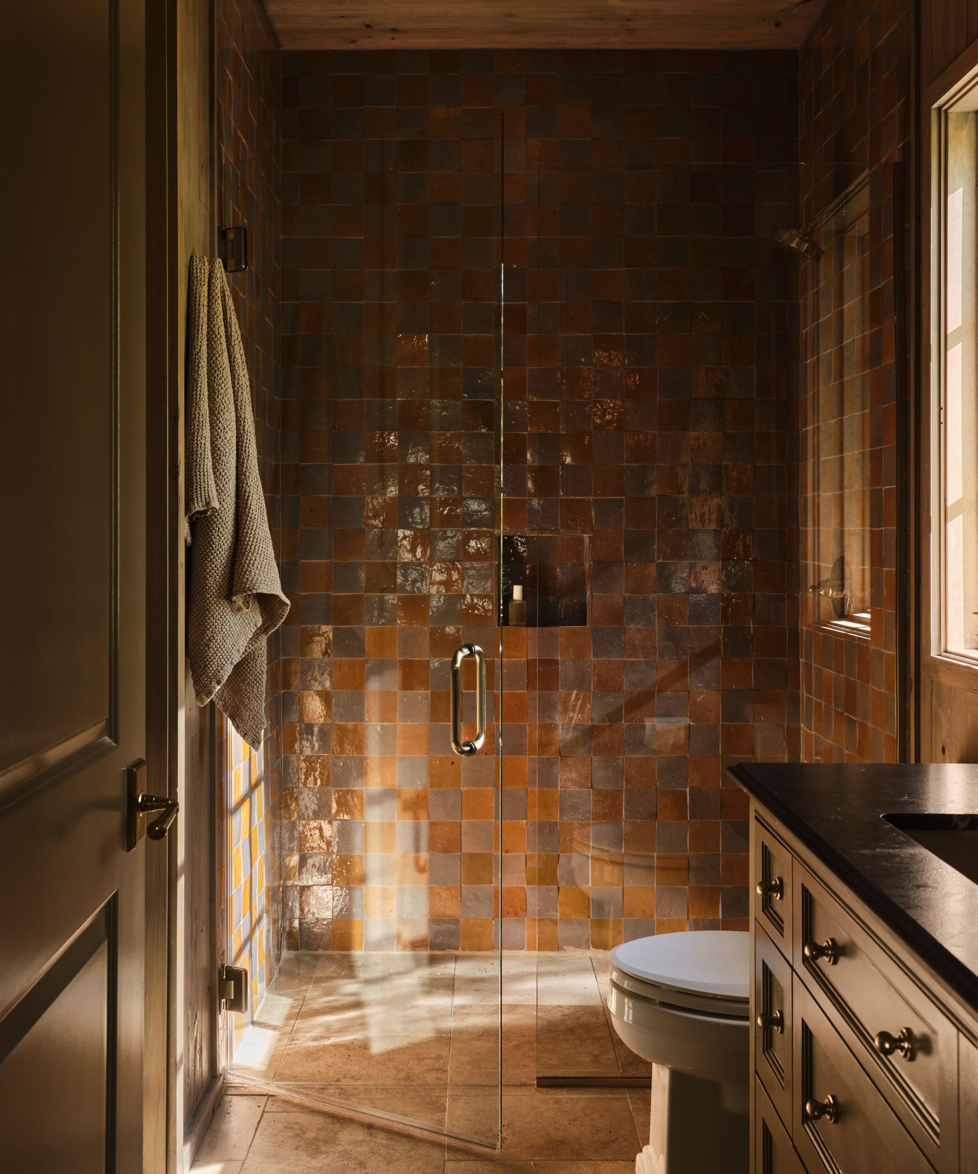 a moody toffee colored bathroom with a shower tiled with zellige and an earthy green painted vanity