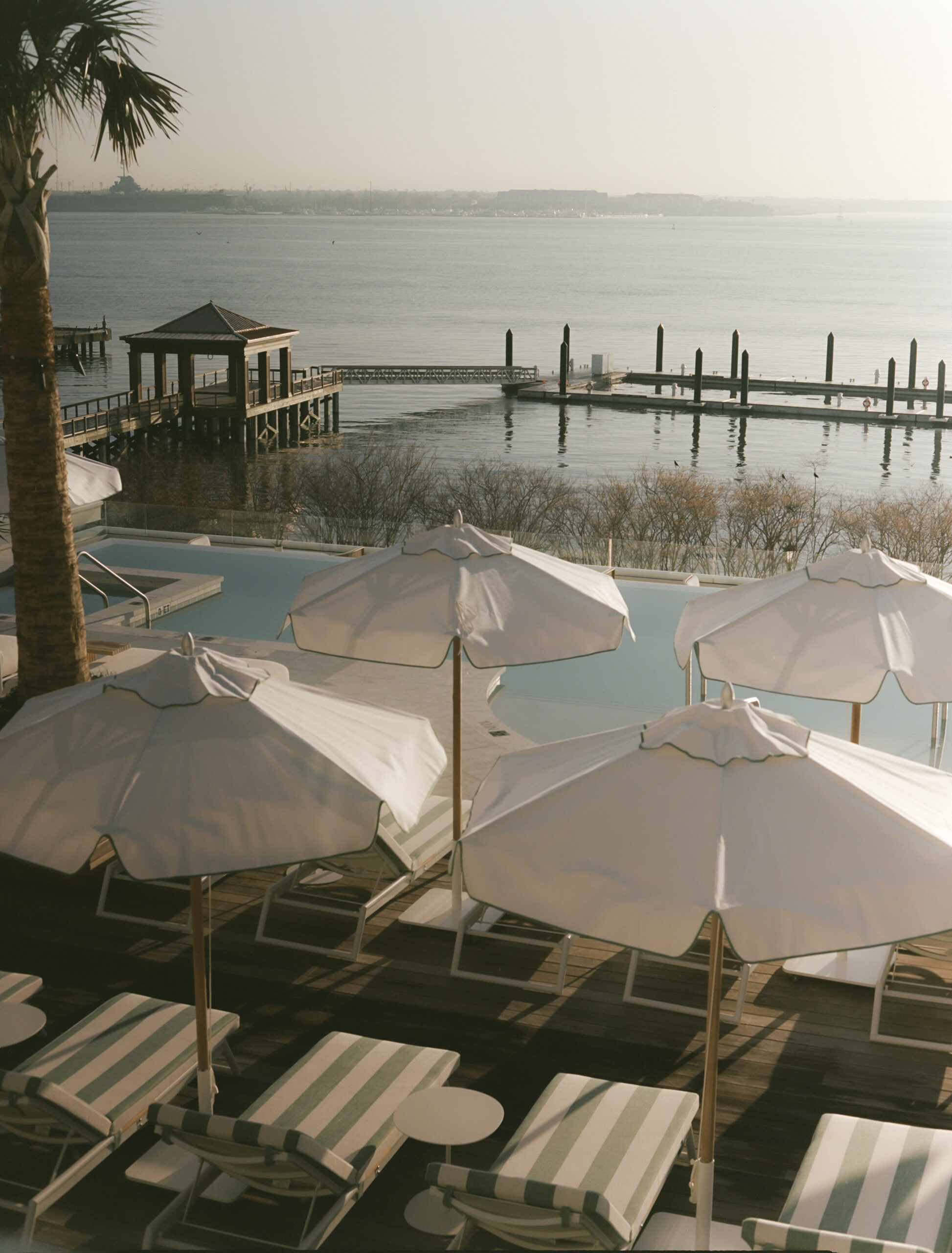 A sun-kissed infinity pool with stripy loungers, white parasols, and dark wood overlooks a calm sea at dawn.