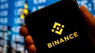 A phone held in someone's hand close to the camera shows the Binance logo, while stock numbers are seen on a blurred screen in the background