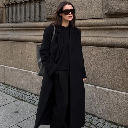 Annabel wears a black long coat, black sweater, and black trousers.