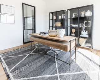 Modern home office featuring a dark beige central desk with drawers on gray area rug, glass front cabinet with minimal against the wall