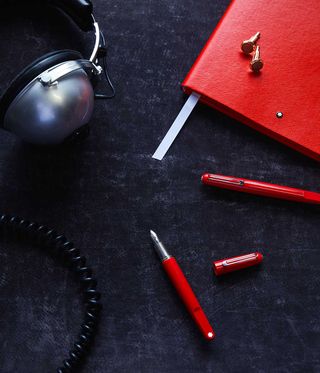 Black wash desk top, edge of silver headphone and black spiral wire, comer of red binder with white ribbon tag pulled out & two gold cufflinks sat on top, closed red fountain pen next to an open red fountain open with lid placed to the right