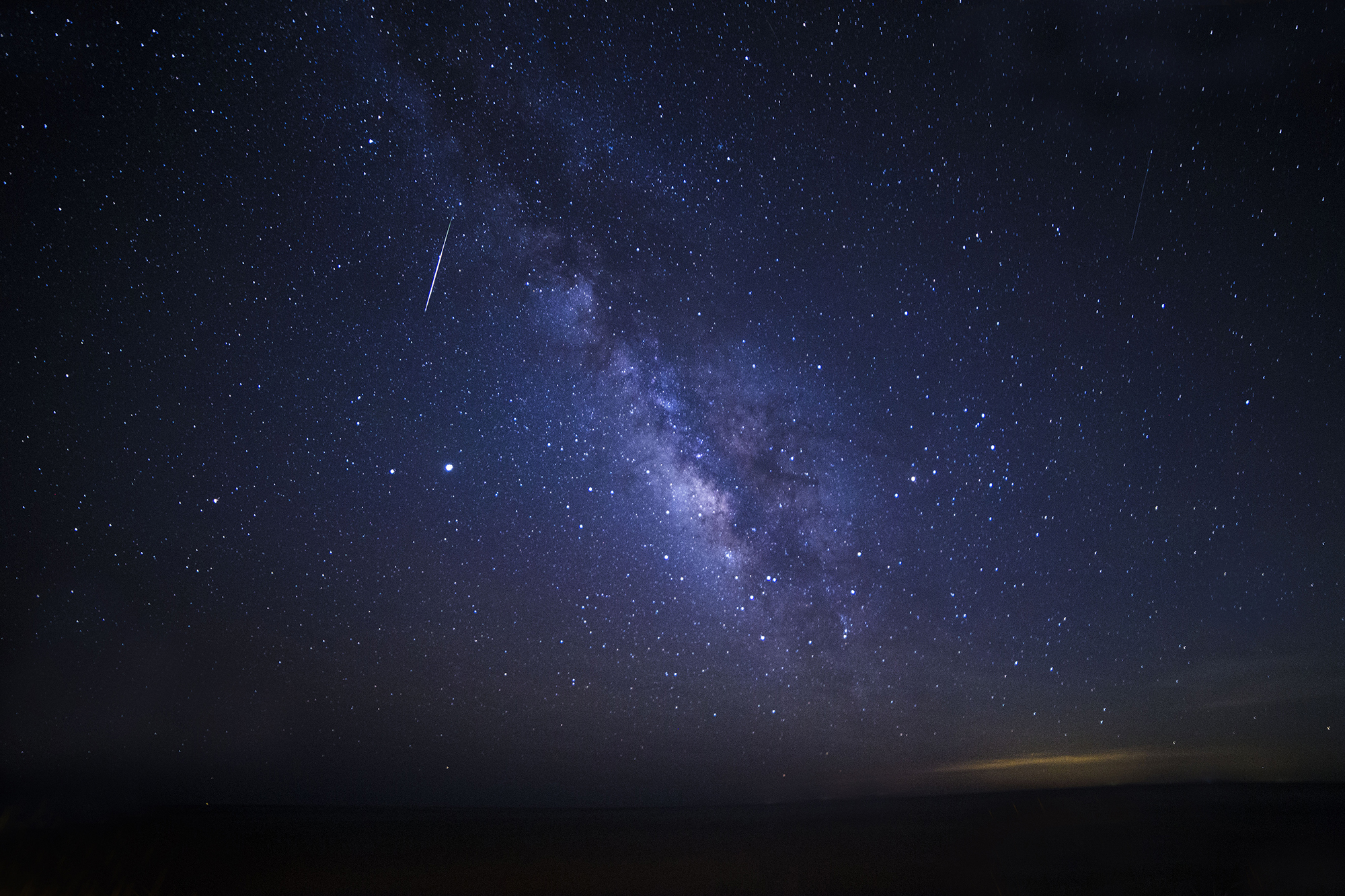 milky way streaks across the center of the image and a meteor in the top left corner leaves a long white trail in the sky.