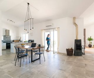 large open plan living room and kitchen with stone tiled floor, dining table, log burner and woman stood at open patio doors