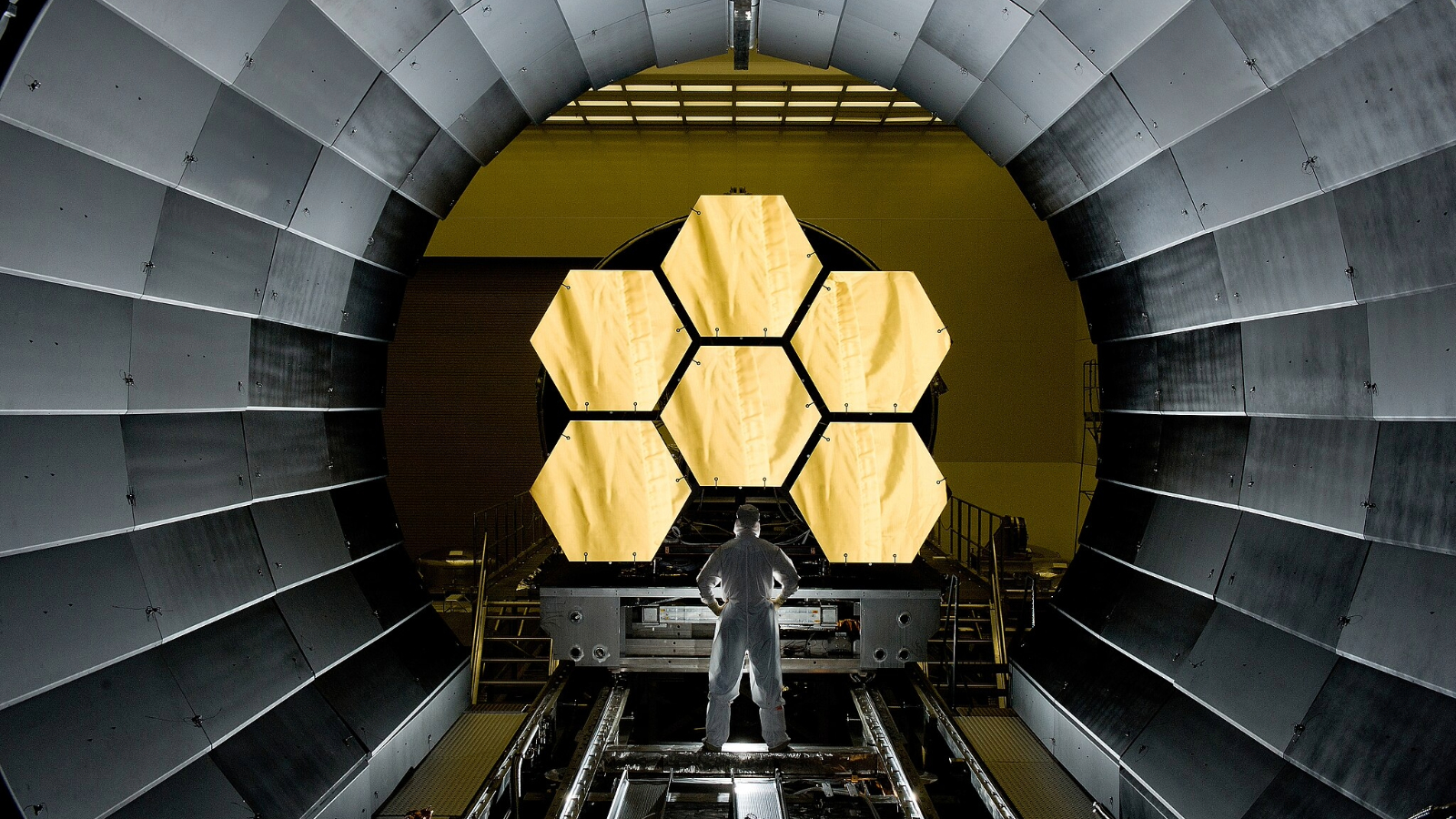 A person in a white outfit stands in front of the James Webb Space Telescope's primary golden mirrors