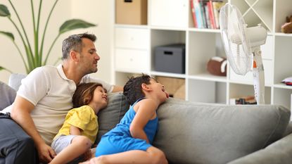 A dad and two kids sit on a couch in front of a fan.