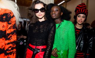 Models are seen wearing patchy knitted jumpers, in black and green. One wears a bright red pair of leather trousers with a black belt.