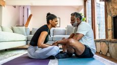 Man and woman sit cross-legged on floor of living room facing each other. 