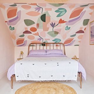 loft bedroom with metal framed bedstead, lilac bedding and a striking sloped ceiling covered in bold graphic floral wallpaper