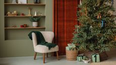 A lit christmas tree with oresents in front of a red curtain with sage green walls, open shelves, white boucle chair with wooden legs and green throw