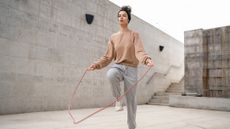 A woman is skipping rope outside. Her hair is tied up and she wears a headband, sweater, sweat paints and sneakers. Behind her is a large concrete wall and a flight of steps.