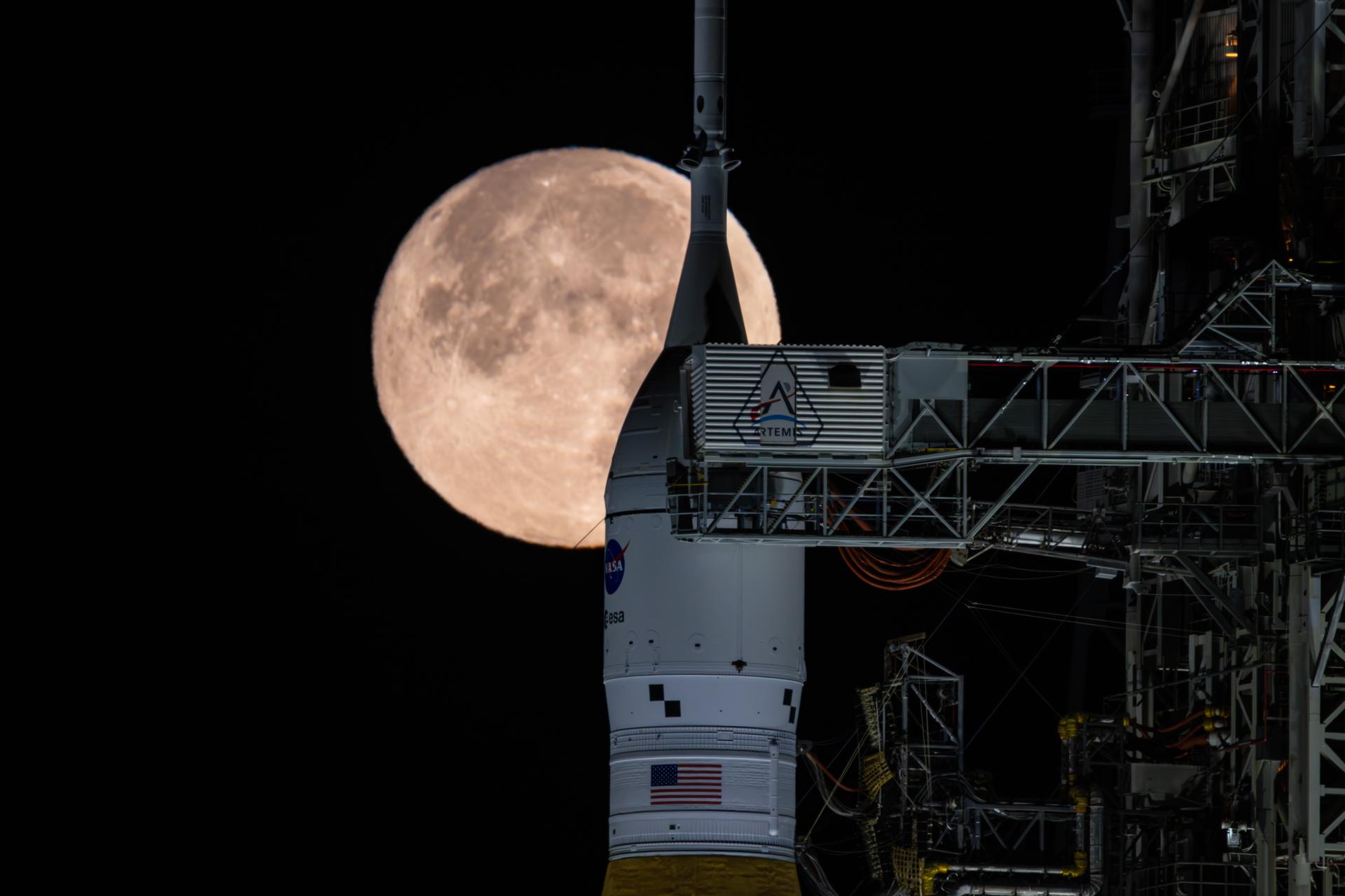 The tip of NASA's Space Launch System rocket is visible with the full moon rising behind it in the black night sky. The steel framework of the launch tower dominates the right of the screen, and the crew arm is extended, bridging the gap to the rocket's white upper stage.