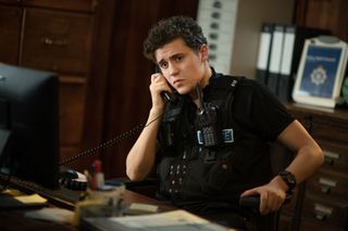 Dylan Llewellyn in character as PC Kelby Hartford. He is sitting in his desk at the police station and answering a phone call.