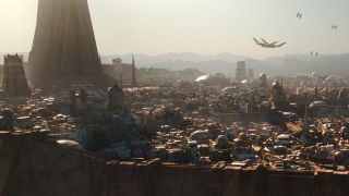 Still from Star Wars media. Here we see a view of the city of Jedha skyline. It is a walled in city with dence buildings in an arid, sandy landscape.