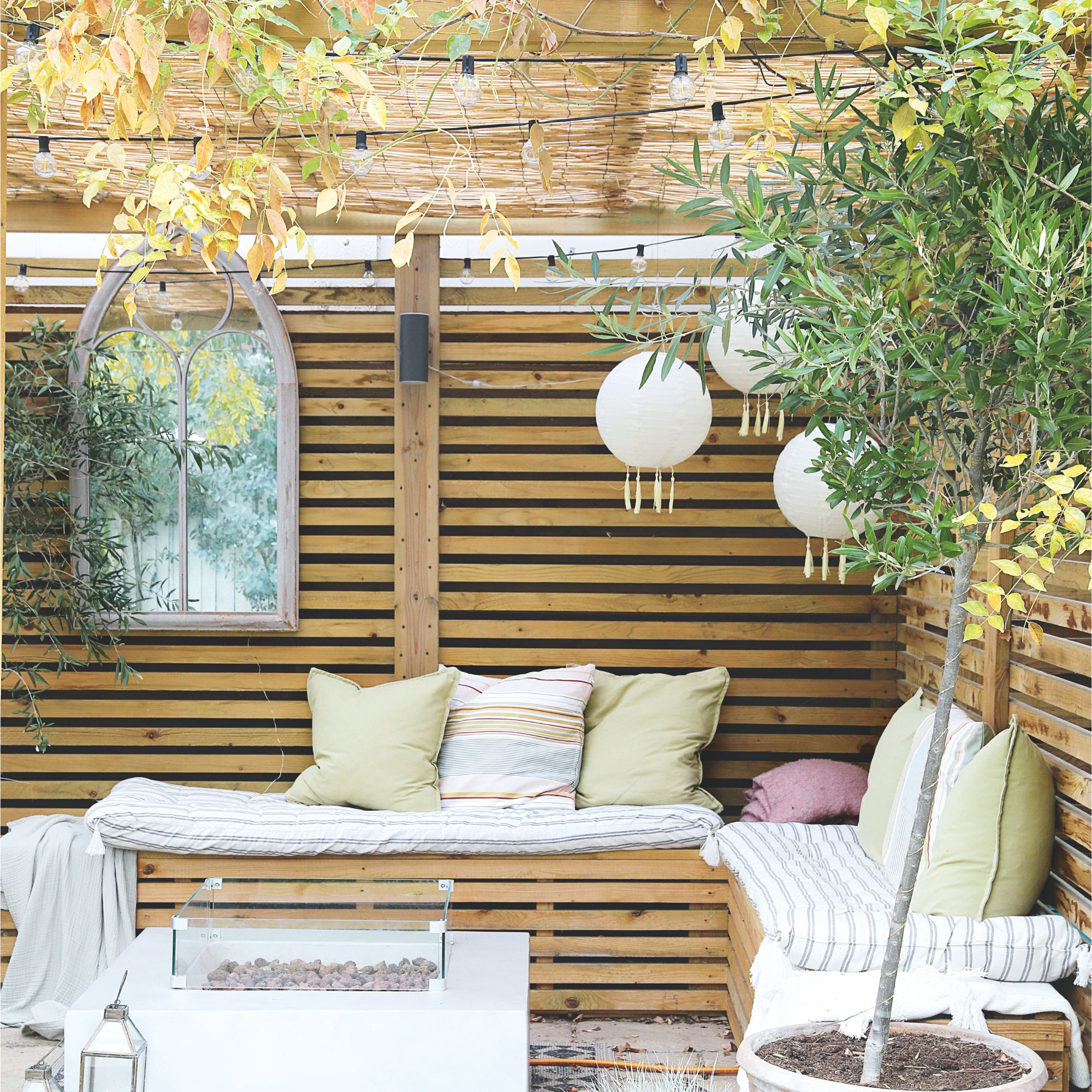 Outdoor seating area with cushions beneath wooden pergola with festoon lights and hanging lanterns
