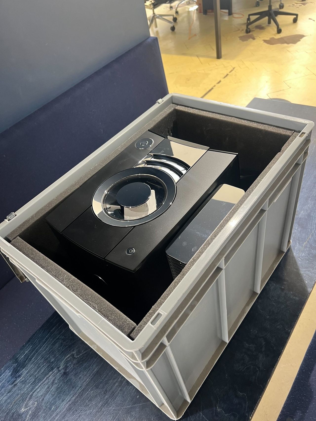 A light gray crate on a black table as seen from above. It's open to reveal a black and silver coffee machine.