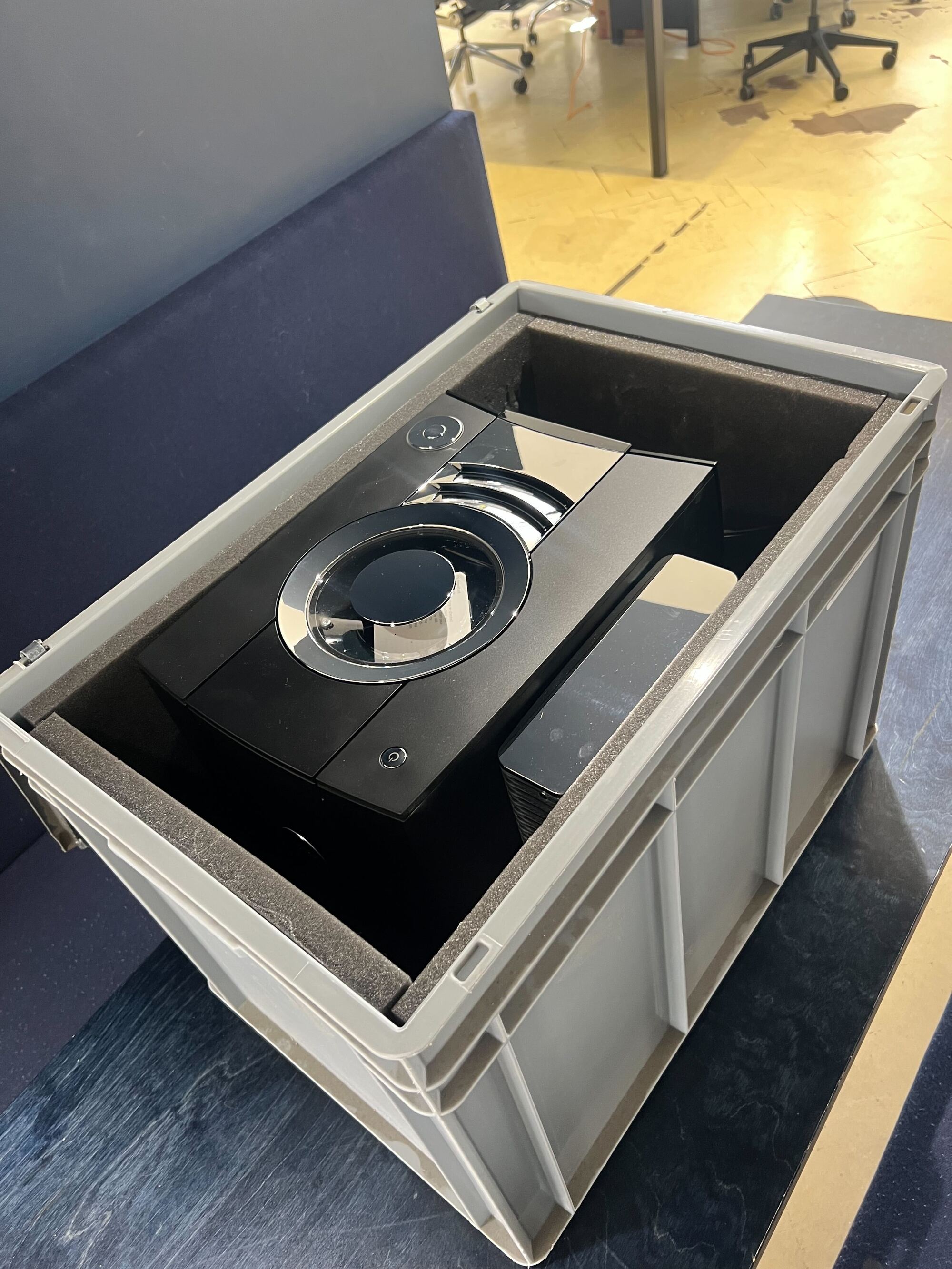 A light gray crate on a black table as seen from above. It's open to reveal a black and silver coffee machine.