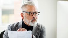 An older man looks serious as he looks at a computer monitor and paperwork.