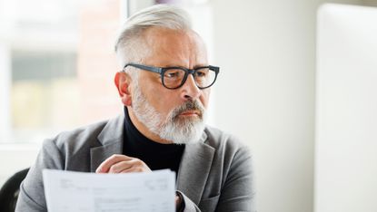An older man looks serious as he looks at a computer monitor and paperwork.