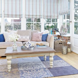 white conservatory with light wooden floorboards, blue and brown striped curtains, cream couch with blue, cream and pink cushions, blue pattern rug under wooden coffee table