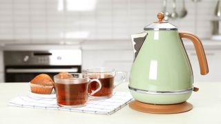 A green electric kettle next to two cups of tea and cupcakes in a kitchen