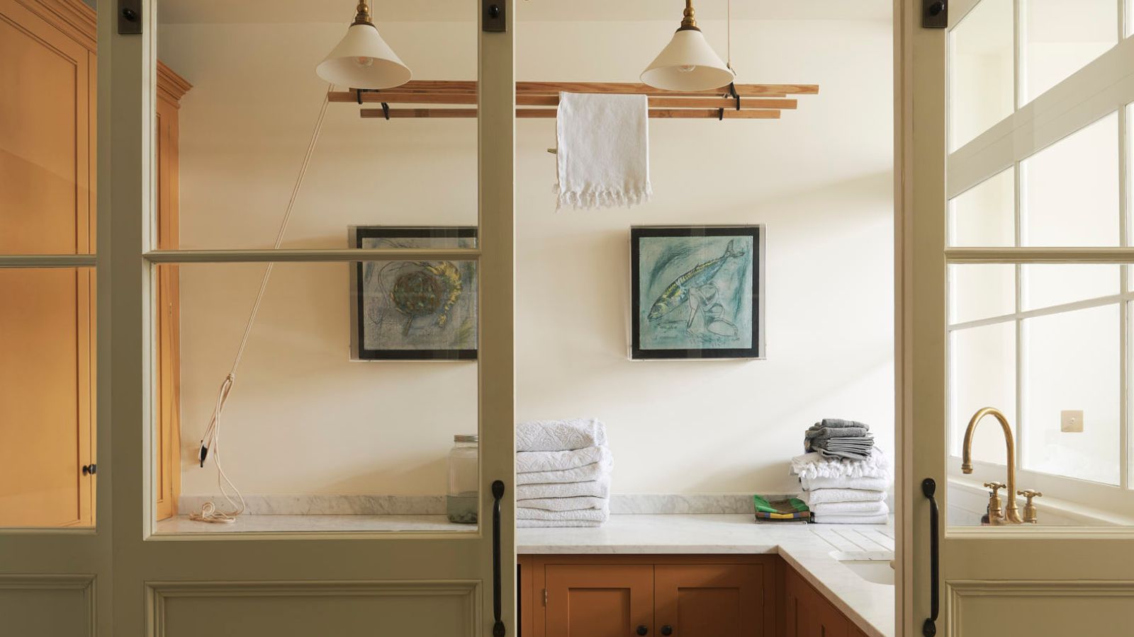 Wooden and cream laundry room with hanging dryer and glass sliding doors. The space has lower cabinets only with a mid-century wooden finish, white marble counters with short upstand, and there are folded towels visible. 