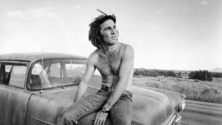 A shirtless Dennis Wilson sitting on the bonnet of a car staring into the distance