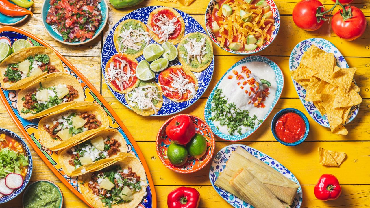 Spread of mexican food on a yellow table, including chiles en nogada, tacos al pastor, chalupas pozole, tamales, chicken with mole poblano sauce