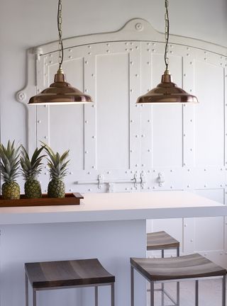 A white kitchen with kitchen island, three backless bar stools, three pineapples on table and two metal Davey Lighting School Lights
