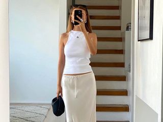 female fashion influencer Orlaith Melia poses for a mirror selfie wearing chunky silver jewelry, a high-neck white tank top, mini black bag, and cream drawstring slip skirt