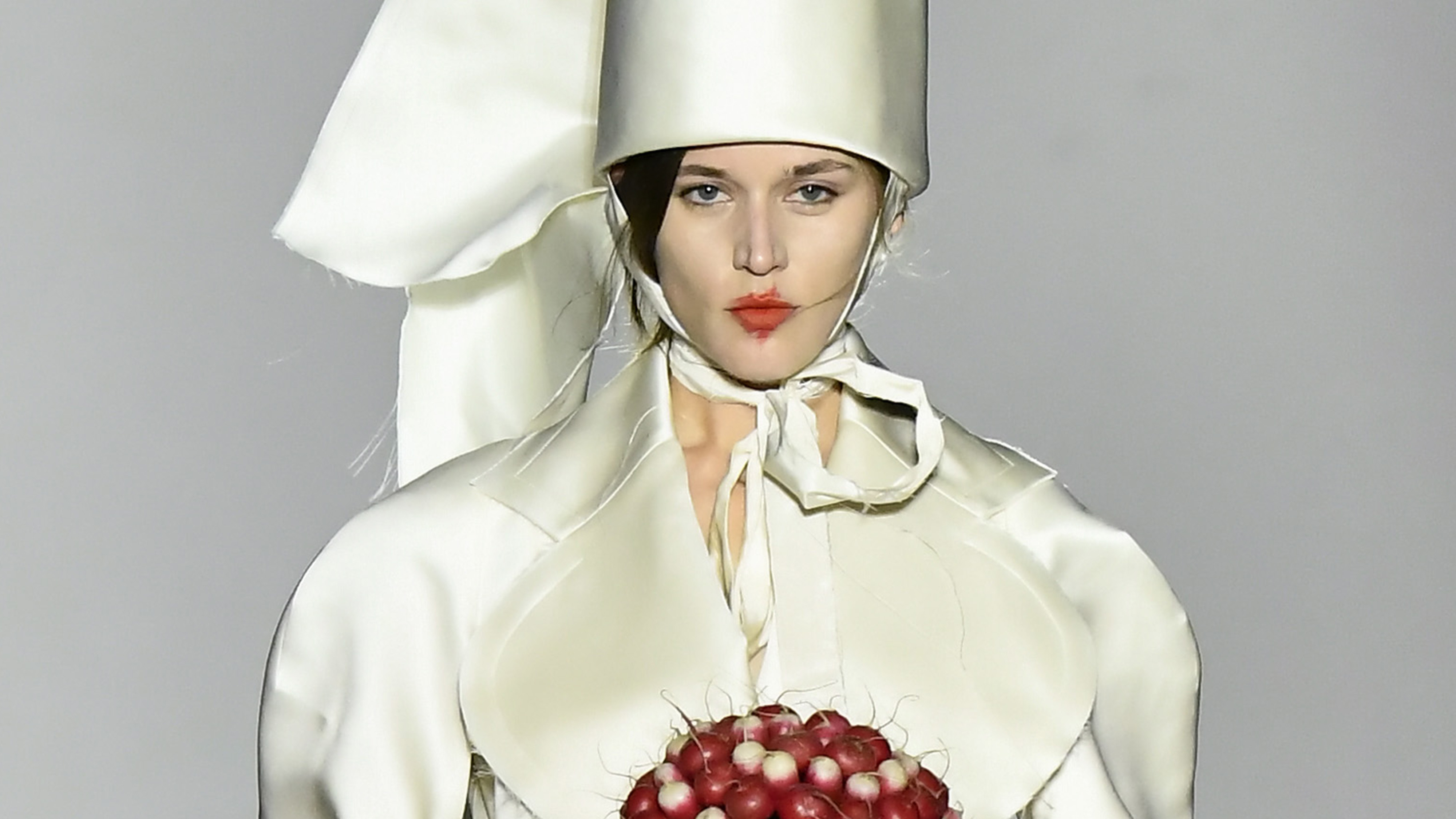 a model wears a tall ivory hat, an ivory blazer, and carries a radish bouquet.
