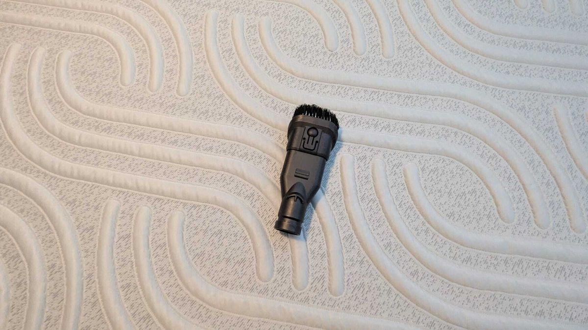 The image shows an upholstery vacuum attachment placed on a white mattress