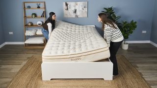 Two women stand at either end of a Saatva Classic Mattress helping to install it on a white bed frame in our sleep studio. With white glove delivery, you can avoid this task