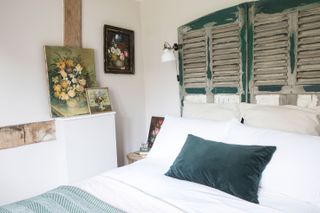 bedhead with faded shutters above bed and green cushion and floral paintings white walls with beams