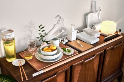 A wooden sideboard with a series of objects by Muji part of a holiday home essentials kit. Including plates and bowls, a jug, a light and scent diffuser, hangers, pen and notebook, towels, and broom and dustpan