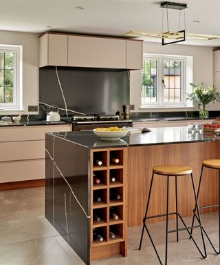 A black marble waterfall kitchen island with a brown base with a wine shelf, two wooden stools, and light pink cabinets and windows behind it