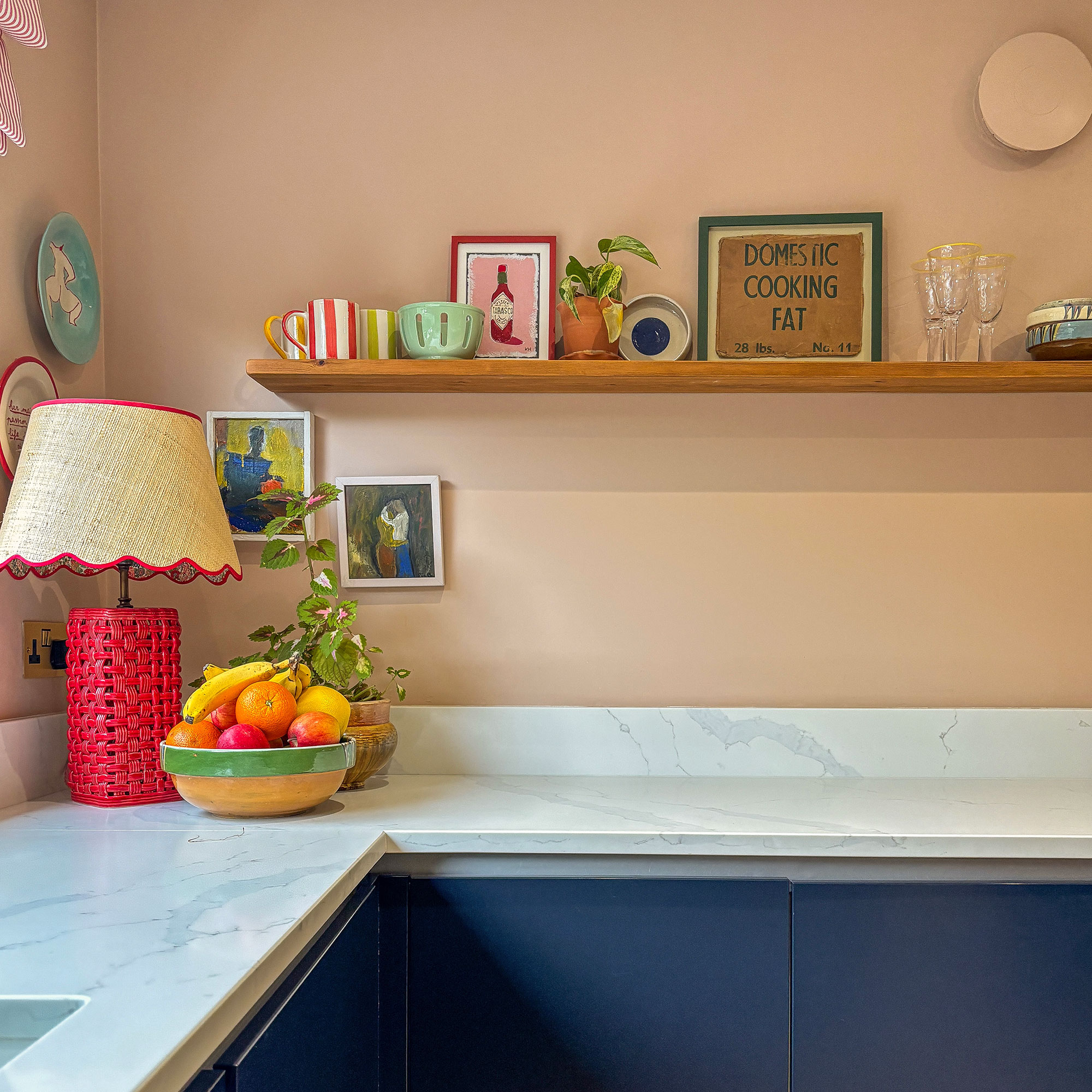 Peach kitchen with red lamp, fruit bowl, art on walls and floating shelf with artwork and drinking vessels above stone worktop and blue cabinetry