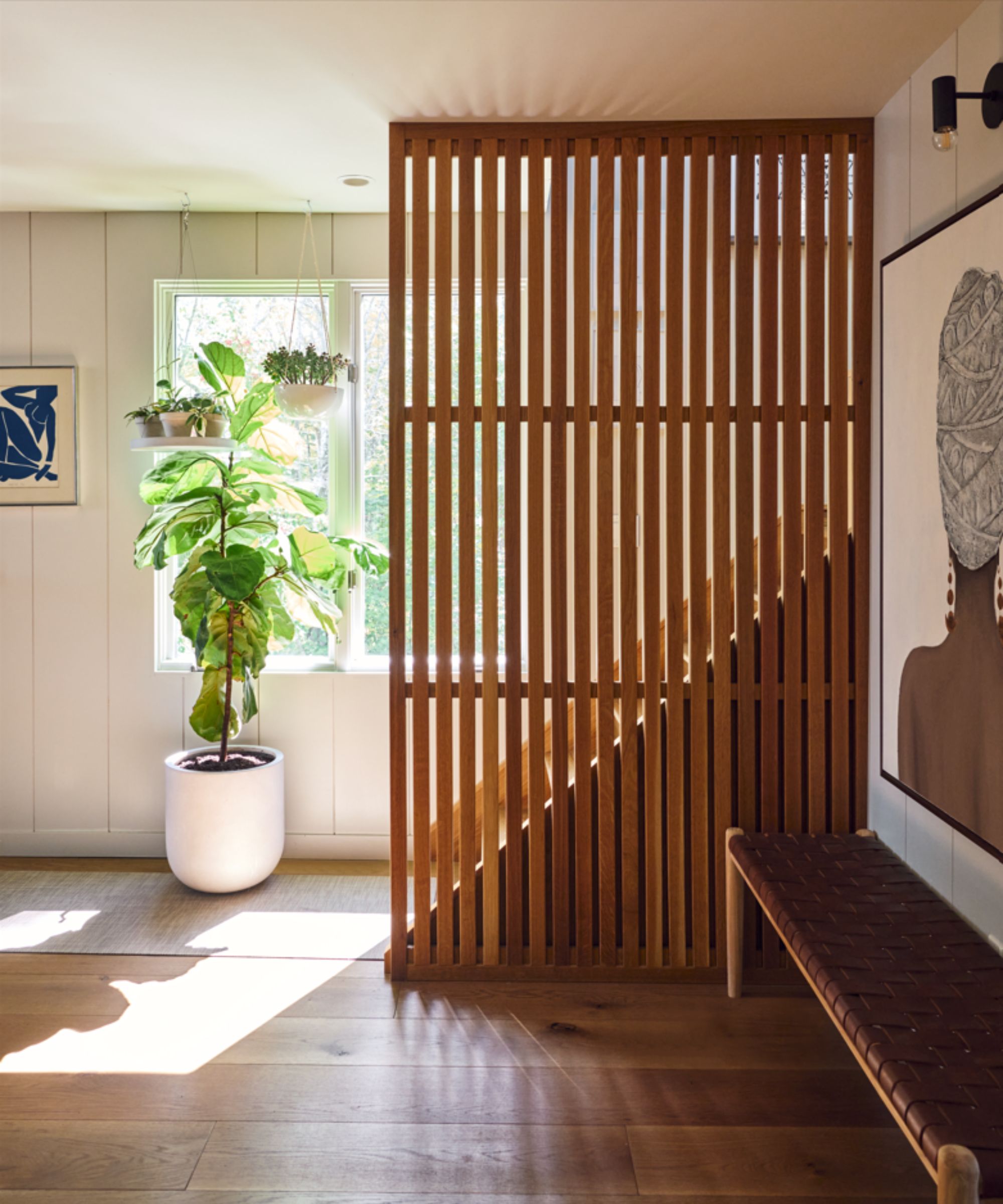 Hallway with sunlight streaming through window, wooden panelled screen in front of a set of stairs, planter by window, warm wooden floor, white panelled walls, artwork and planters