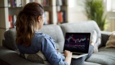 A young woman looks at a trading graph on her laptop while sitting on her sofa.