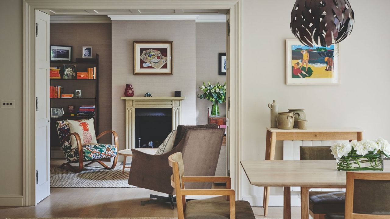 A living room dining room with beige walls, a fireplace, eclectic vases and flowers, a black bookcase filled with orange books and framed artwork on the walls